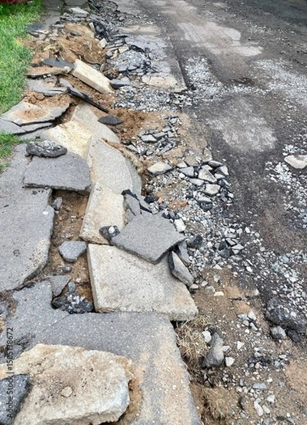 Fototapeta Damaged road surface with broken asphalt and gravel. Uneven pavement and debris scattered along the roadside. Signs of construction or repair work in progress.