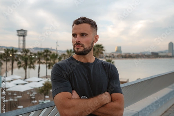 Fototapeta Portrait of a businessman posing with crossed arms in Barcelona, Spain, with beach and cityscape in background
