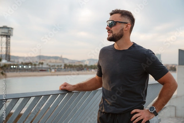 Fototapeta Sportsman wearing sunglasses, relaxing after training, gazing at the beautiful Barceloneta beach and the shimmering sea in Barcelona