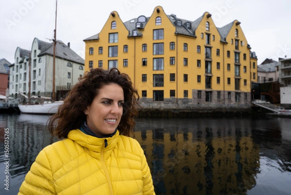 Fototapeta Portrait of a young brunette woman looking away, wearing a yellow anorak, with the Brosundet Canal and singular building in Alesund, Norway, behind her