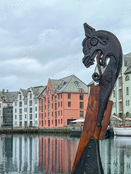 Fototapeta Viking figurehead of a ship prow ornament docked in the Brosundet canal, with colorful Art Nouveau buildings of Alesund, Norway reflected in the calm water