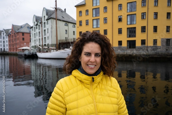 Fototapeta Portrait of a young brunette woman looking at the camera, wrapped in a yellow down jacket, with the Brosundet Canal in Alesund, Norway, behind her, with art nouveau buildings