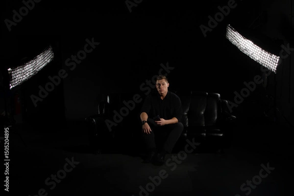 Fototapeta Man Sits on Black Couch Under Studio Lights Holding Phone