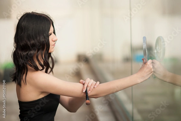 Obraz An attractive Young Woman in a black dress touches a makeup brush to her arm. She is posing in a modern city, and her reflection is visible in a glass wall