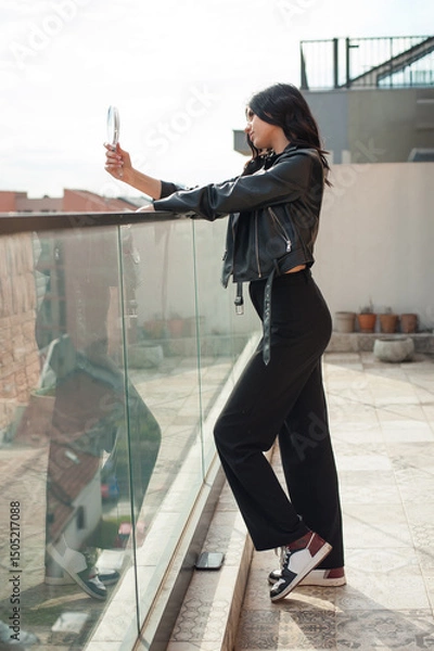 Obraz A young, fashionable woman is posing near a glass barrier, looking at herself in a handheld mirror. She is on a rooftop during the daytime, wearing black clothing