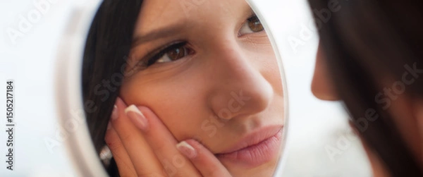 Obraz A young woman looks at her reflection in a mirror, gently touching her face with her hand. She is inside. The natural light suggests it is daytime. Her nails are manicured