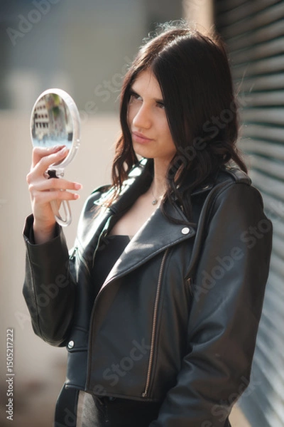 Obraz A young woman with dark hair, wearing a stylish black leather jacket, stands outdoors. She holds a round, handheld mirror and attentively examines her reflection