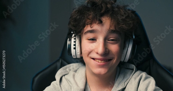 Fototapeta A boy with curly long hair and a sweatshirt sits in a chair wearing wireless headphones. He focuses on the computer screen during a quiet evening at home.