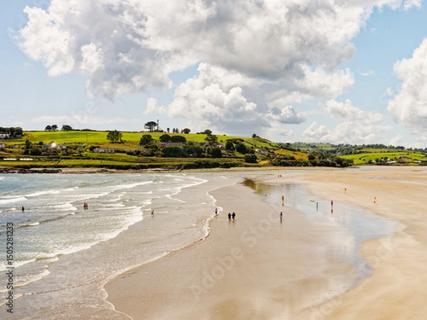 Fototapeta West Cork, June 25, 2023. A stunning view of a beach, with people enjoying a summer's day. The contrast between the sand, sea and green hills creates a peaceful, beautiful scene.