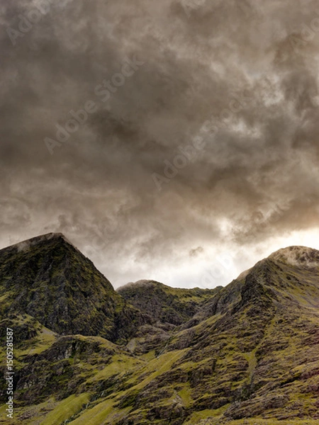 Fototapeta Carrauntoohil, the highest mountain in Ireland, is surrounded by swirling clouds as dusk falls. The landscape is full of green hills and rocky outcrops.