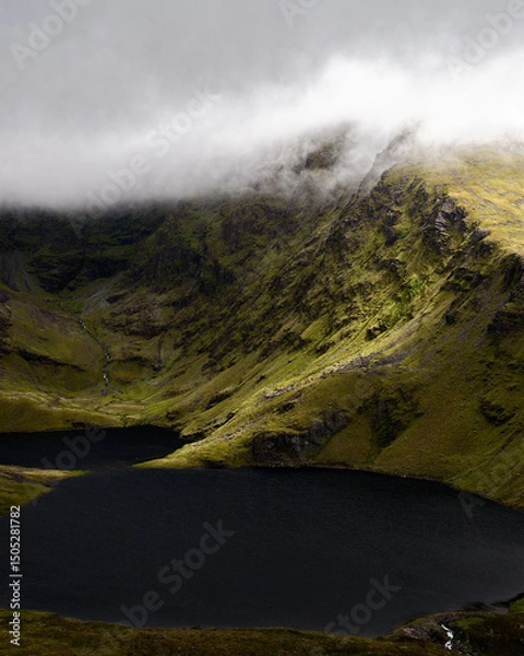 Fototapeta Jagged peaks pierce through swirling mist above a calm, dark lake. The scene conveys feelings of mystery and awe-inspiring natural beauty.