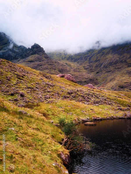 Fototapeta Lough Callee is nestled among rocky hills and cliffs under a moody sky. The landscape is covered in green and brown grasses, with a few small trees growing near the water's edge.