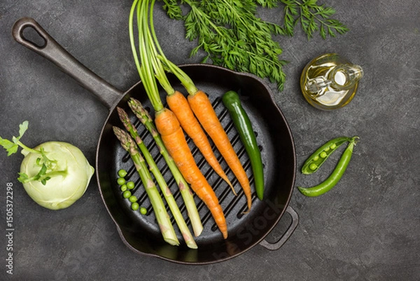 Fototapeta Fresh carrots with green tops, green pepper pod and asparagus in frying pan.