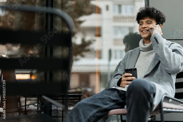 Fototapeta Young man talking on his cell phone while sitting in a chair with a coffee cup in his hand in front of a window