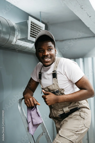 Fototapeta Worker smiling while on ladder, preparing for home maintenance in a cozy indoor space during daylight hours