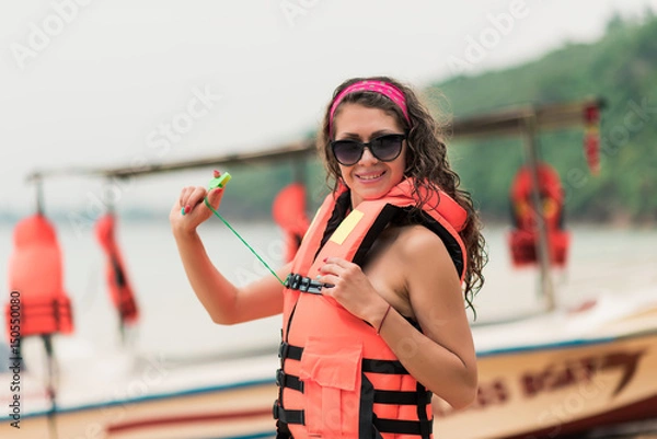 Fototapeta A girl on the beach in a life jacket and glasses looking at the camera and smile