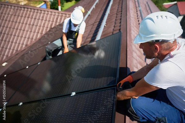 Fototapeta Workers building solar panel system on rooftop of house. Two men installers in helmets installing photovoltaic solar module outdoors. Alternative, green and renewable energy generation concept.