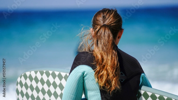 Fototapeta A woman wearing a wetsuit holding a patterned surfboard looks out towards the ocean. The blue water and clear sky create a serene and adventurous seaside atmosphere at this popular Cornish resort.