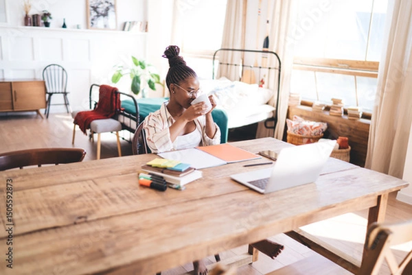 Fototapeta Woman working remotely from stylish home interior, reaching toward laptop with one hand while sitting at rustic desk filled with planners, markers, phone and digital accessories