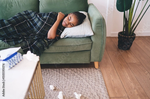 Fototapeta African American woman lying sick on green couch under blanket, eyes closed and resting with tissues scattered on floor, showing fatigue and need for recovery