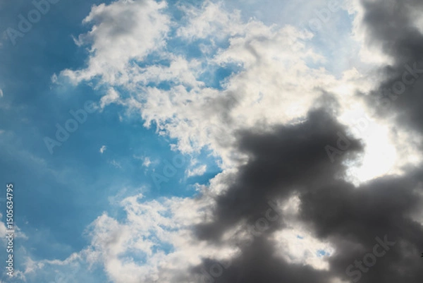 Fototapeta The sun hidden behind a mass of dark grey clouds, with rays of light and shadows on blue sky, for background