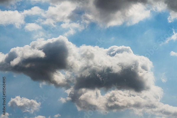 Fototapeta The sun hidden behind a mass of dark grey clouds, with rays of light and shadows on blue sky, for background