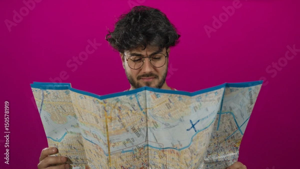 Fototapeta Young man with beard and glasses studying a map against a vibrant pink background, portraying curiosity and exploration.