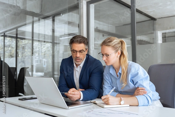 Fototapeta Two partners, team of professional business people working, discussing, writing notes at desk. Mature focused senior mentor leader man showing young woman manager project on laptop in modern office