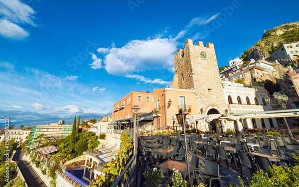 Obraz Stone clock tower and empty outdoor terrace in Taormina old town, Sicily. Bright morning sunlight, traditional Mediterranean architecture and scenic hillside view create classic Taormina atmosphere