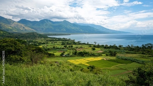 Obraz Expansive panoramic view of a large tranquil lake bordered by vibrant green agricultural fields and distant majestic mountains under a cloudy sky.