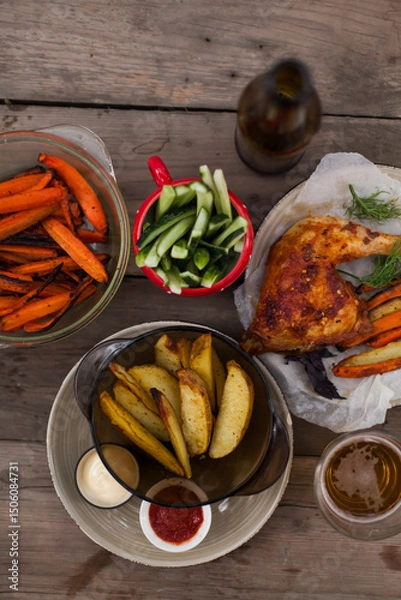 Fototapeta Top-down view of pub food: baked chicken drumsticks, rustic fries, sliced pickles, sauces, and beer. Cozy and casual atmosphere. Perfect for menus, blogs, or food delivery visuals