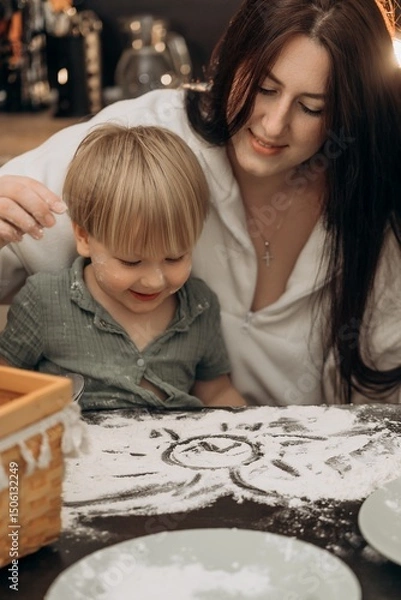 Fototapeta Mother and toddler smiling while drawing a sun in flour on the table during a cozy kitchen moment filled with love, messy play, warmth, and gentle parenting in a natural home setting