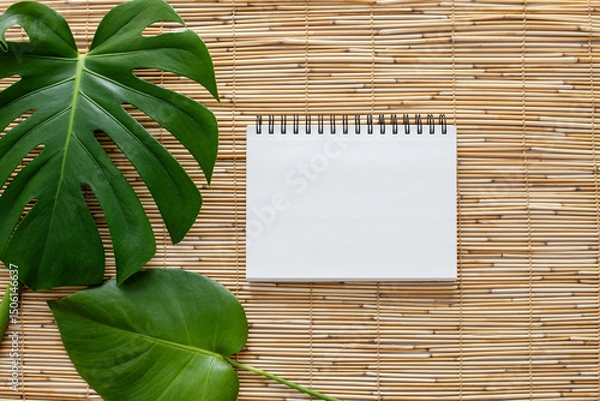 Obraz Top view flatlay of a blank white page of spirallled notebook on straw reed shade suface with two green monstera leaves