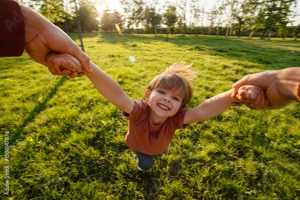 Obraz Portrait of a happy boy holding hands and spinning in a circle with his parent, having fun in a park on sunset. A carefree happy childhood. 