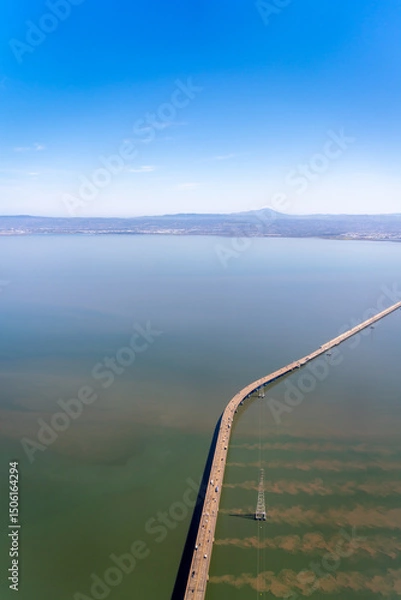 Fototapeta Aerial view of a long bridge stretching across a body of water under a clear blue sky.