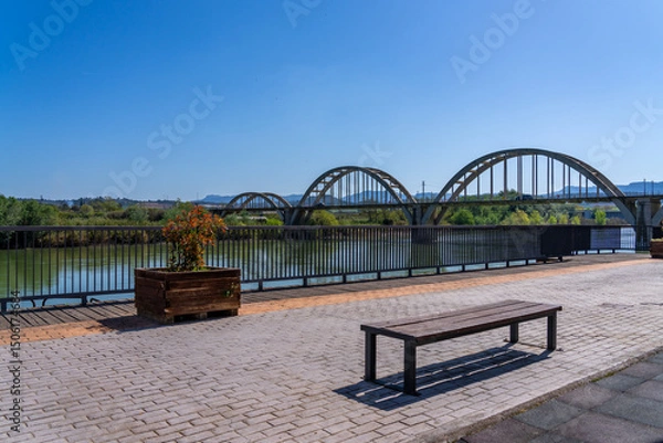 Fototapeta Scenic view of a bridge over a river with a bench and planter in the foreground.
