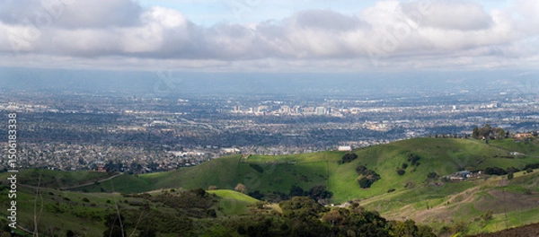 Fototapeta A panoramic view of a city from rolling green hills under a cloudy sky.