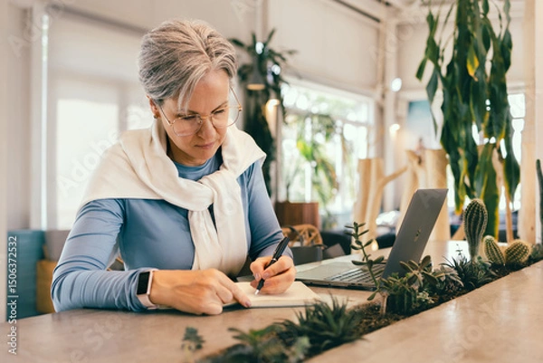 Fototapeta Focused senior woman with gray hair writing in notebook while working on laptop in cozy modern cafe
