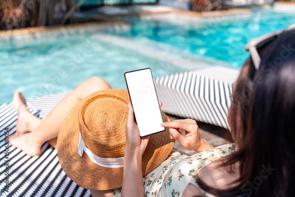 Fototapeta Young woman tourist using smart phone at resort pool while traveling for summer vacation