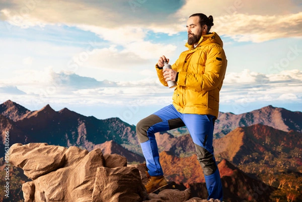 Obraz Young man explorer climbing the mountain range and hydrating with his water bottle, drinking refreshment on top of the rocky mountain. Male hiker refreshing after the trekking activity.