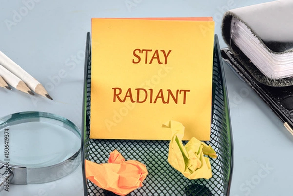 Fototapeta A bright yellow sticky note with the message stay radiant sits in a black mesh organizer at a desk. Crumpled pieces of paper and office tools create a busy yet inspiring workspace environment