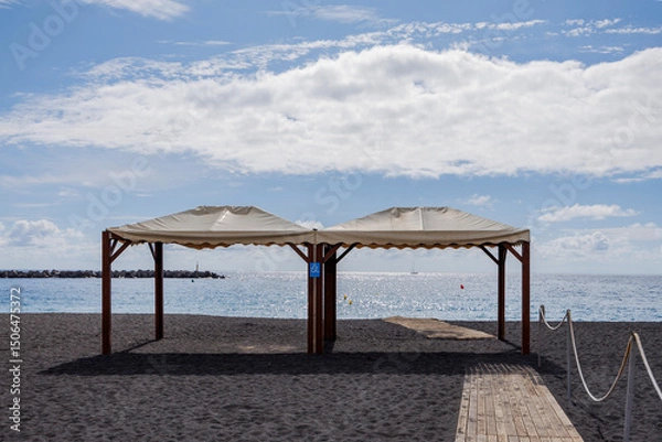 Fototapeta Symmetrical beach scene with a wooden path leading to an empty canopy by the sea, expressing the serene stillness before summer begins.