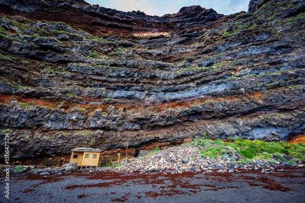Fototapeta Small wooden structure nestled against dramatic layered volcanic cliffs and surrounded by black sand, illustrating scale, solitude and natural resilience.