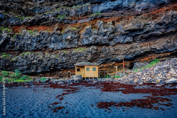 Fototapeta Small wooden structure nestled against dramatic layered volcanic cliffs and surrounded by black sand, illustrating scale, solitude and natural resilience.