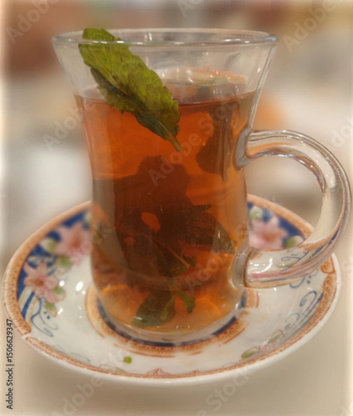 Fototapeta A close-up view of herbal mint tea with fresh mint leaves, presented in a clear glass on an ornate floral-patterned saucer.