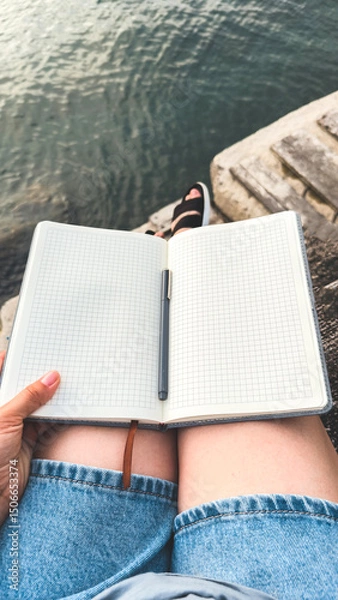 Fototapeta A person women sitting by the water with an open squared notebook on their lap, holding a pen, wearing denim shorts and sandals, relaxing on stone steps near a calm body of water. vertical