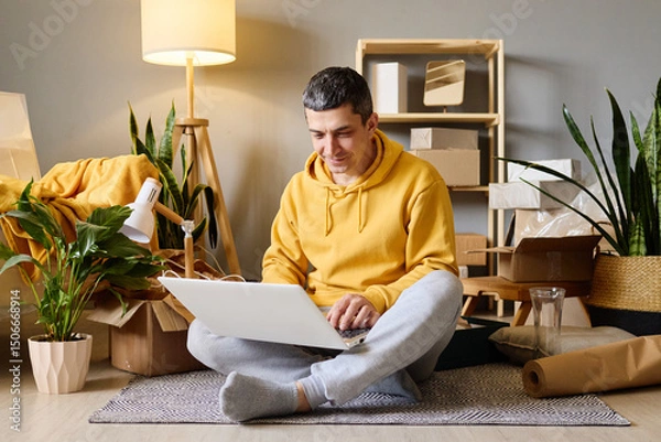 Fototapeta A man sits on the floor among moving boxes, using his laptop after moving into his new home.