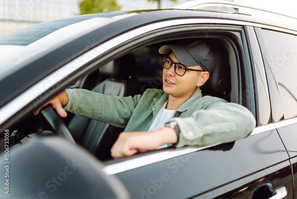 Obraz Young man sitting behind the wheel of the car, enjoying the sunny weather, looking out the window. Stylish car driver with an open window. Transport, car travel concept.
