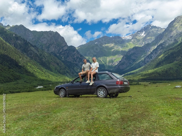 Obraz Adventurous couple sitting on the roof of their car, taking in the breathtaking panorama of a lush green valley nestled amidst towering mountains, enjoying a memorable road trip