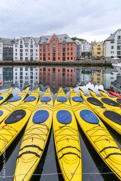 Fototapeta Bright yellow kayaks moored on the calm waters of Brosundet canal in Alesund, Norway, with colorful Art Nouveau buildings reflected in the background on a cloudy day